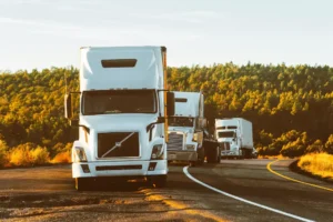 Três caminhões brancos percorrem uma estrada rural cercada por vegetação e floresta. A imagem destaca o papel do transporte rodoviário na logística sustentável, alinhado aos princípios ESG, promovendo eficiência, responsabilidade ambiental e boas práticas na cadeia de suprimentos.