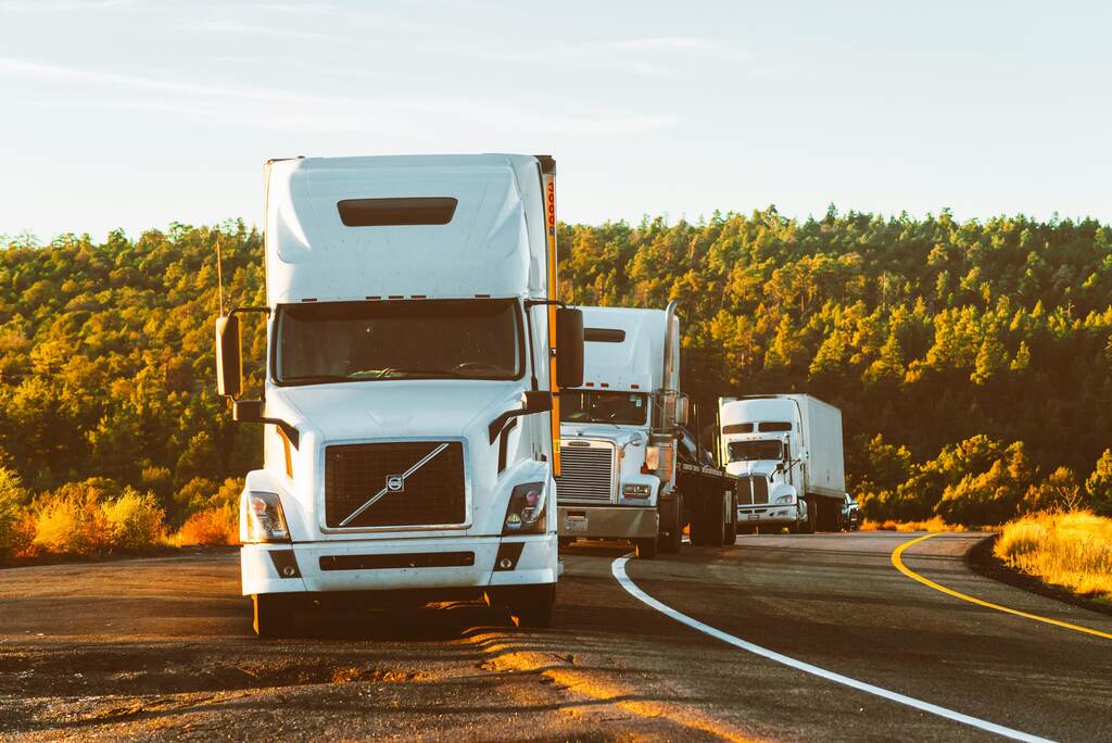 Três caminhões brancos percorrem uma estrada sinuosa cercada por floresta ao entardecer, simbolizando a força da logística do biodiesel no Brasil.