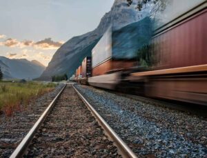 Trem de carga atravessa região montanhosa em alta velocidade, destacando a eficiência da logística do biodiesel no Brasil, que conecta centros produtores a mercados consumidores por trilhos.