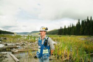 Profissional em campo com capacete e colete de segurança azul, atuando em área úmida cercada por vegetação e troncos. A cena representa atividades ligadas às profissões sustentáveis, como conservação ambiental, manejo de recursos naturais e pesquisa ecológica em ambientes florestais.